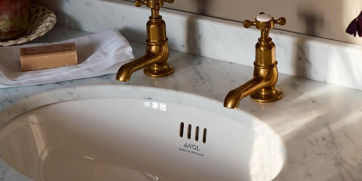 White bathroom sink in a marble countertop with two brass taps and a bar of soap beneath an ornate mirror.