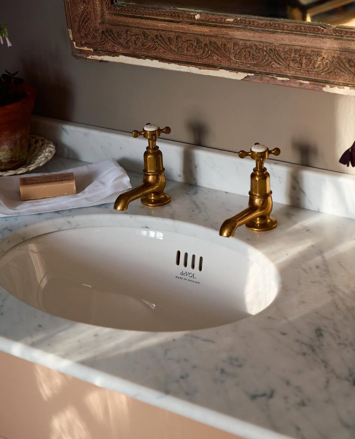 White bathroom sink in a marble countertop with two brass taps and a bar of soap beneath an ornate mirror.