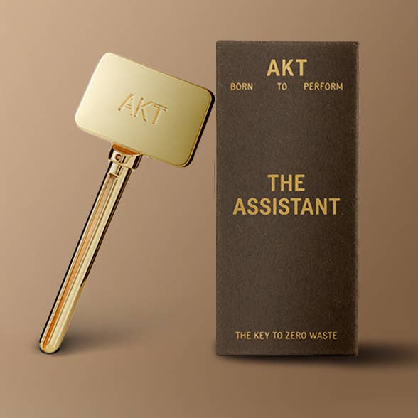 Gold tool with 'AKT' branding next to a brown box labeled 'The Assistant' on a beige background. Deodorant