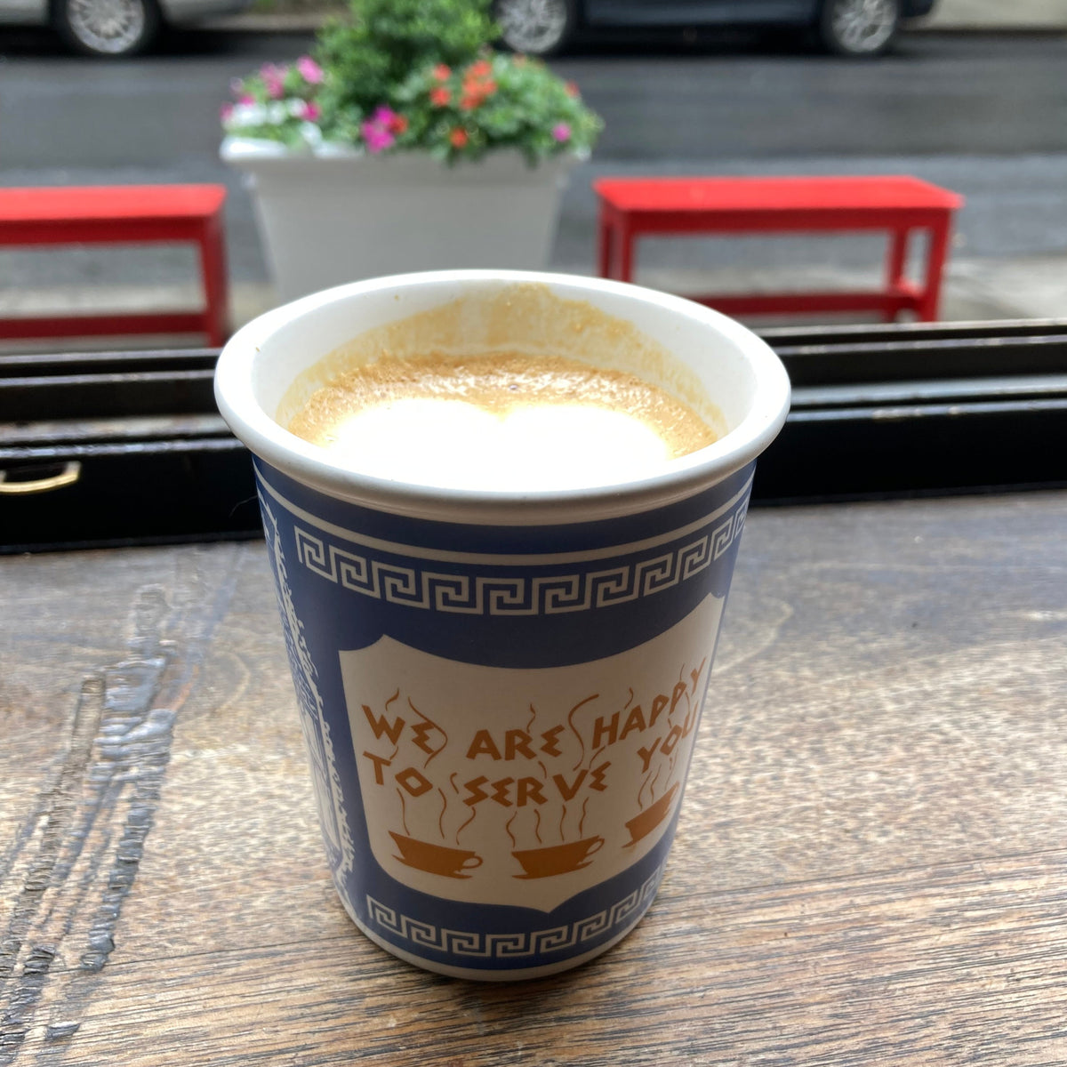 A ceramic We are Happy to Serve You" coffee cup with text on it, placed on a wooden, cafe table. New Yorker's favorite mug.