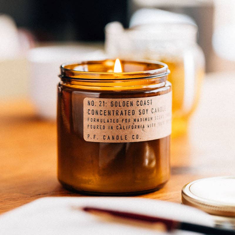Lit P. F. Candle Co. Golden Coast candle in an amber glass jar on a wooden surface.