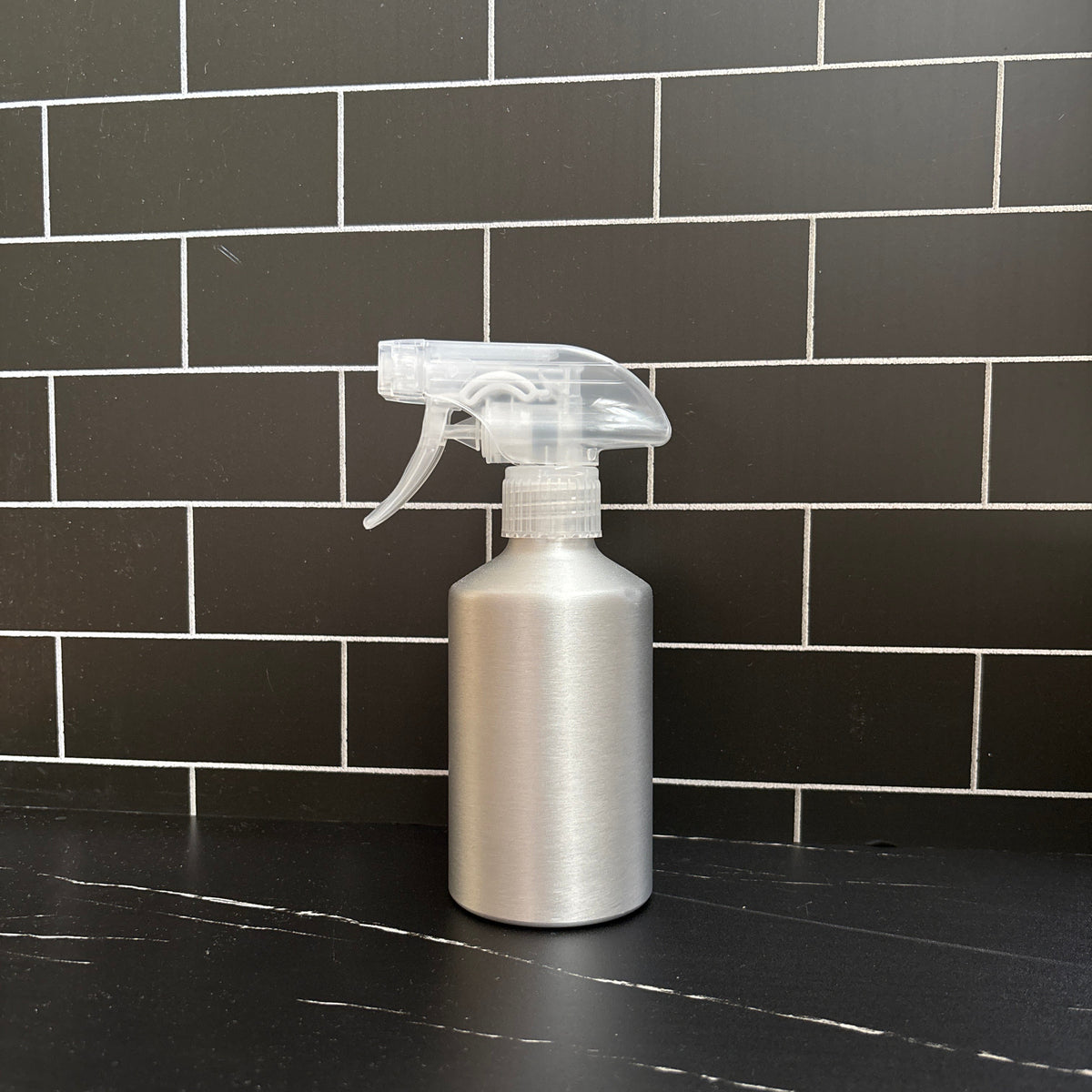 Aluminum spray bottle with clear nozzle against a black tiled wall