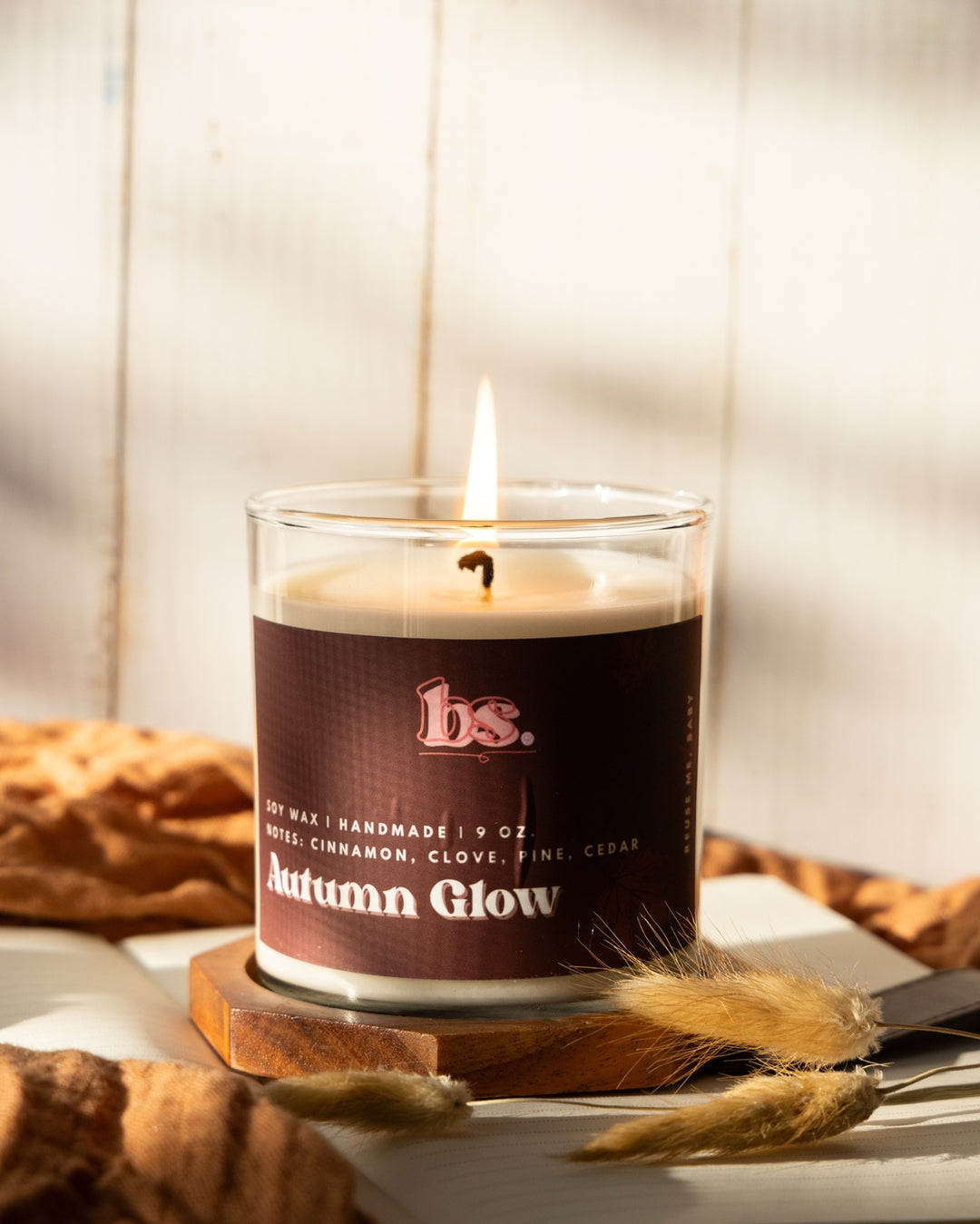 Candle labeled 'Autumn Glow' with a brand logo, placed on a surface with a blurred background.