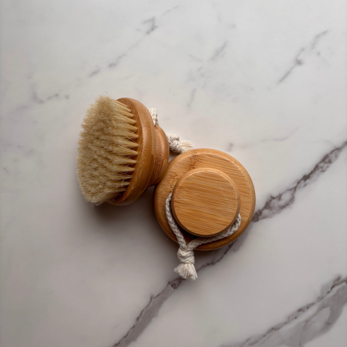 A wooden hand brush with natural bristles and a matching round wooden cap tied together by a white rope, placed on a white marble surface with gray veining.