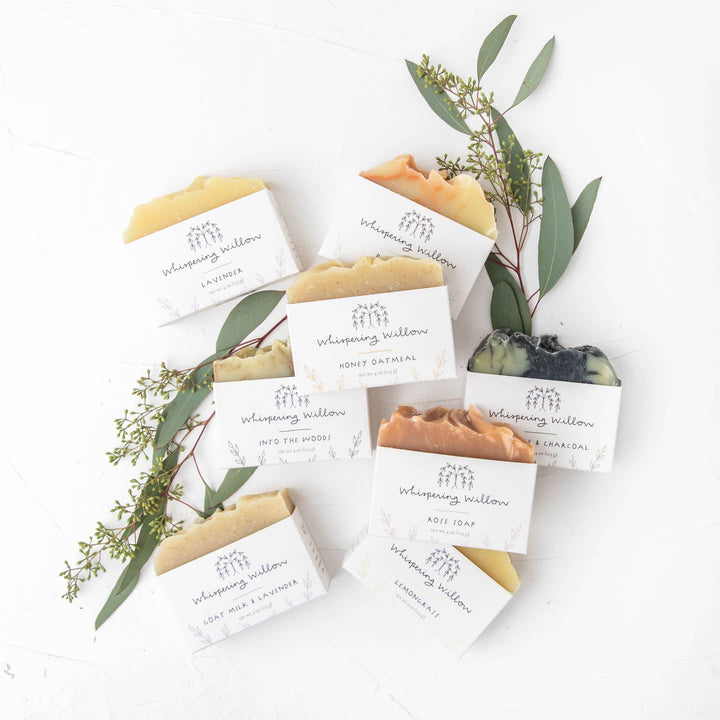 Bar soaps from Whispering Willow with a gentle yellow color, displayed on a white surface with green leaves and packaging.