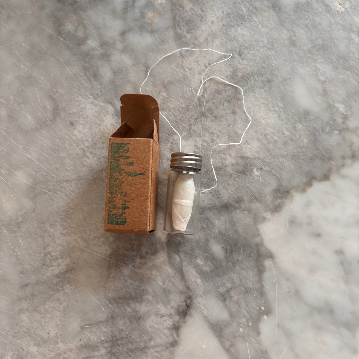 A small glass vial with white powder and a metal cap, placed beside an open brown cardboard box with a green city skyline illustration, on a gray marble surface.