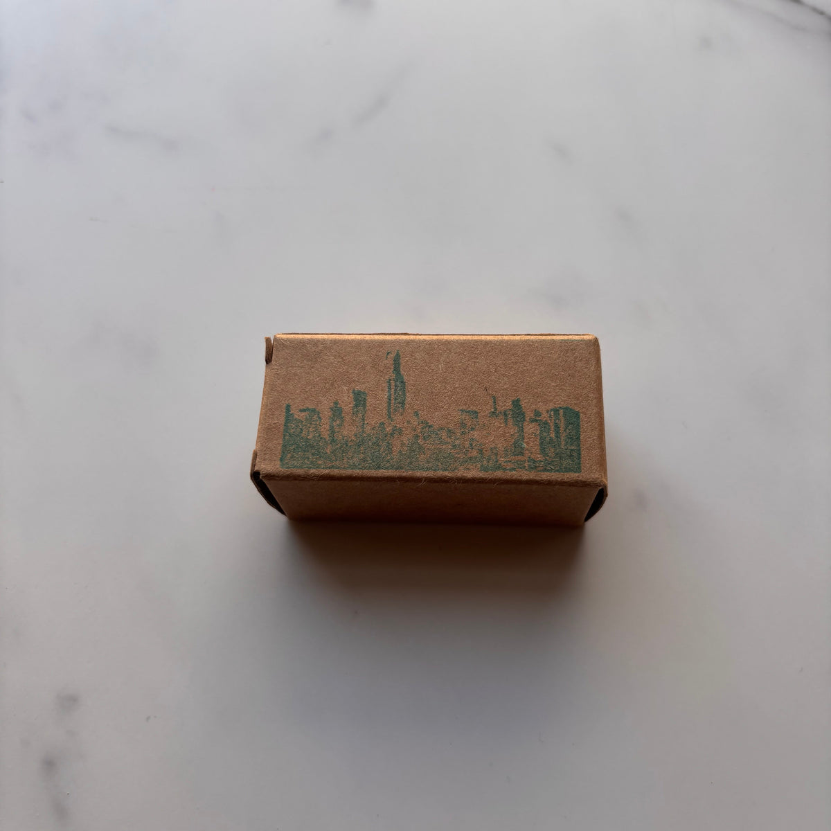 A small brown kraft paper box with a green printed logo, placed on a white marble surface.
