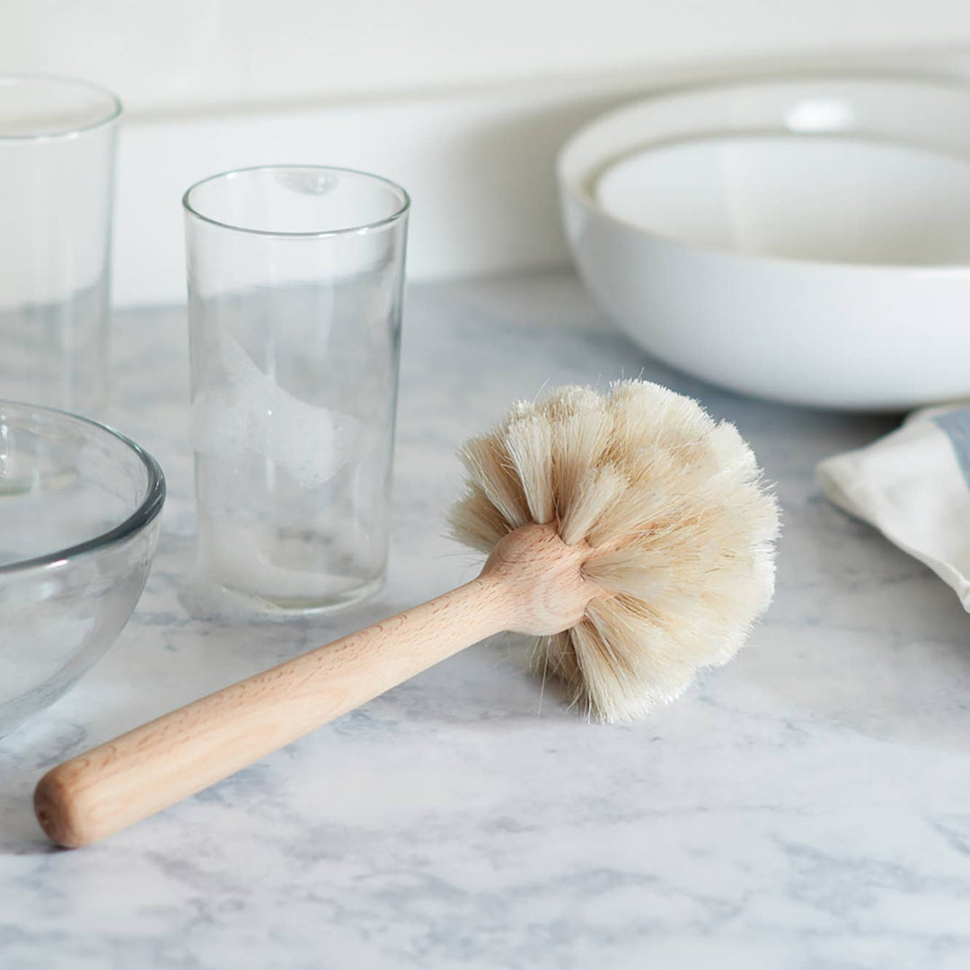 A natural bristle dish brush with a beechwood handle, resting on a marble surface, with a clear glass and a white bowl in the background.