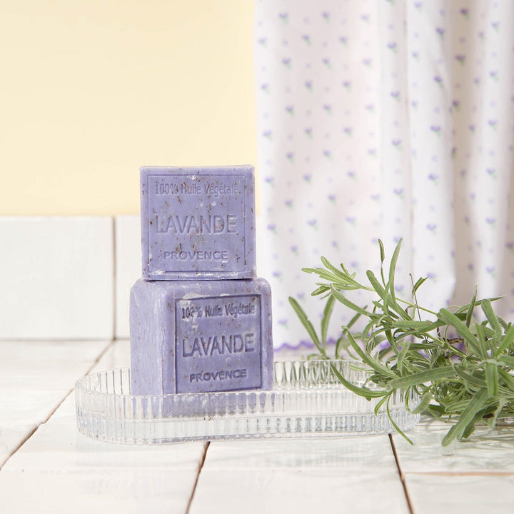 Two bars of lavender soap on a glass tray with a plant in the background.
