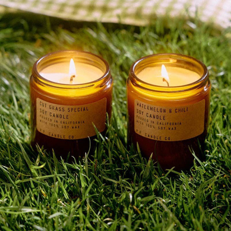 Two lit candles in amber jars on grass, with visible labels.