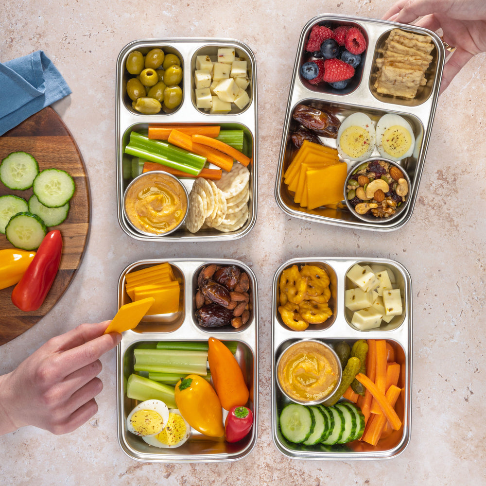 Four metal bento boxes filled with a variety of colorful vegetables and snacks on a light stone surface.