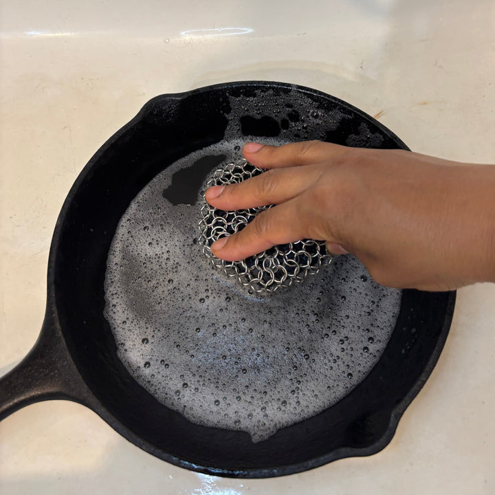 Person scrubbing a black cast iron skillet with a stainless steel chain sponge on a white surface