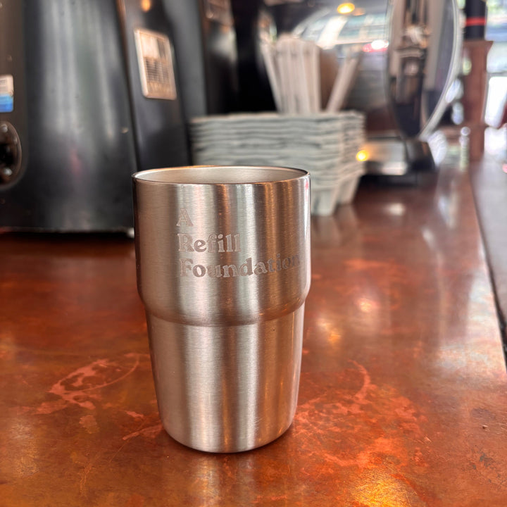 A brushed stainless steel travel tumbler with 'A Refill Foundation' engraved on the side, sitting on a warm copper-colored counter with coffee equipment blurred in the background.