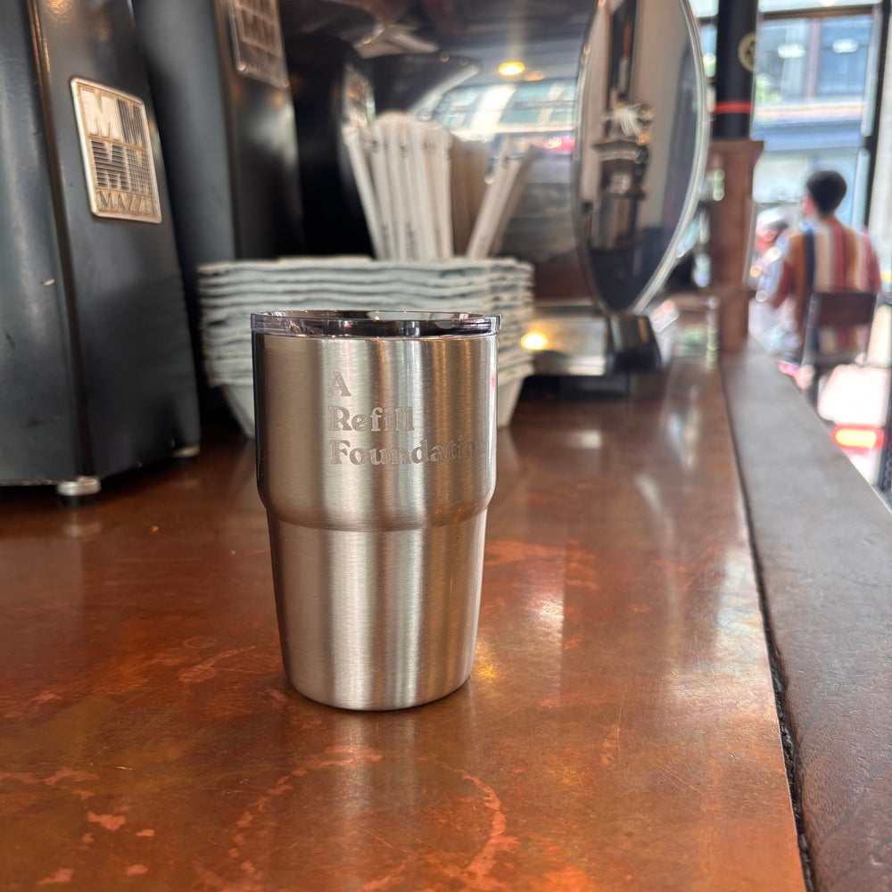 A stainless steel travel tumbler with engraved text reading 'A Refill Foundation' sits on a warm copper-colored countertop in a cafe setting, with coffee equipment blurred in the background.