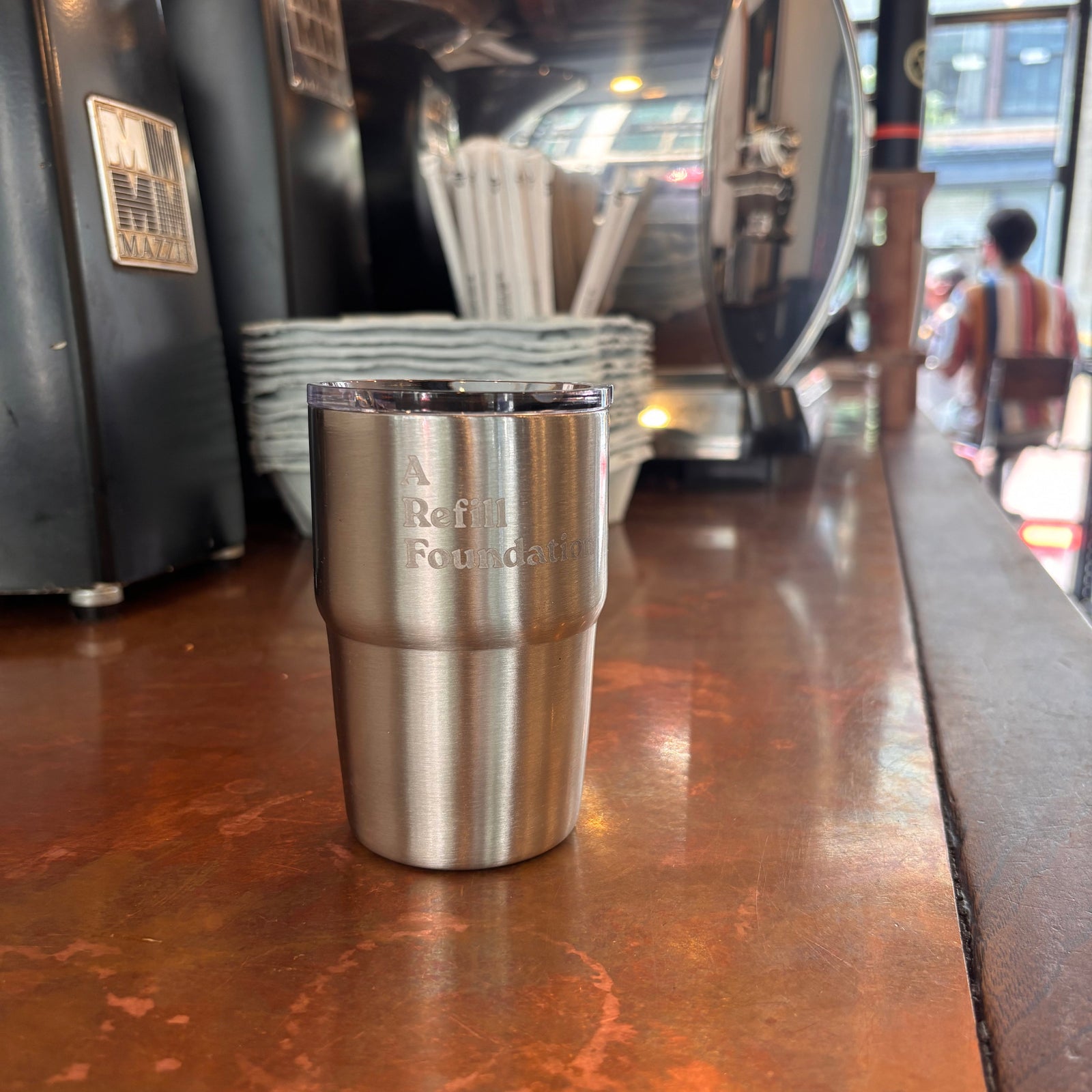 A stainless steel travel tumbler with engraved text reading 'A Refill Foundation' sits on a warm copper-colored countertop in a cafe setting, with coffee equipment blurred in the background.